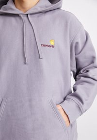 Lavender hoodie made of cotton blend with front pocket, drawstring hood, and embroidered yellow logo. Soft texture and relaxed fit.