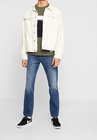 Giacca di jeans bianca sopra una maglietta verde oliva, abbinata a jeans blu e sneakers nere. L'outfit presenta una vestibilità casual e colori a contrasto.