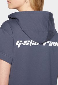 Harmaa huppari, jossa on suuri valkoinen "G-Star Raw" -logo selässä. Varustettu kiristettävällä hupulla ja lyhyillä hihoilla, jotka on valmistettu teksturoidusta kankaasta.