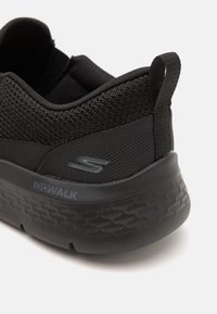 Zapatilla negra con parte superior de malla, acentos texturizados, suela acolchada y un logotipo "GO WALK" en el lateral. Tira en la parte posterior para facilitar el calzado.