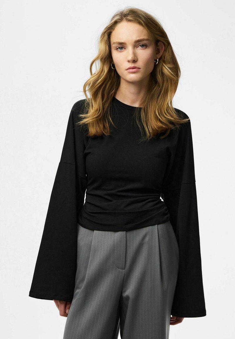 Top nero a maniche lunghe con maniche larghe, realizzato in tessuto morbido, abbinato a pantaloni grigi a righe sottili, caratterizzati da una vestibilità comoda e dettaglio in vita arricciata.