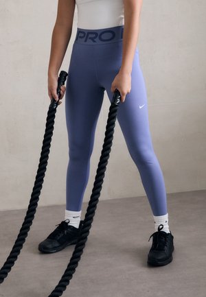 Personne portant des leggings Nike violets, des chaussettes blanches et des chaussures noires tenant des cordes de combat noires dans un espace d'entraînement intérieur.