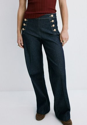 Mujer con jeans oscuros de talle alto y pierna ancha, con botones dorados y una blusa de canalé sin mangas de color óxido.