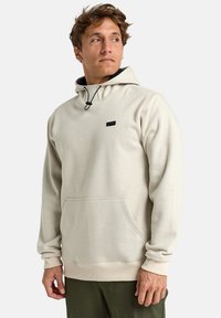 Jasno beżowa bluza z kapturem na sznurku, z przednią kieszenią kangurką, ściągaczami przy rękawach oraz małym czarnym logo na klatce piersiowej. Miękki materiał.