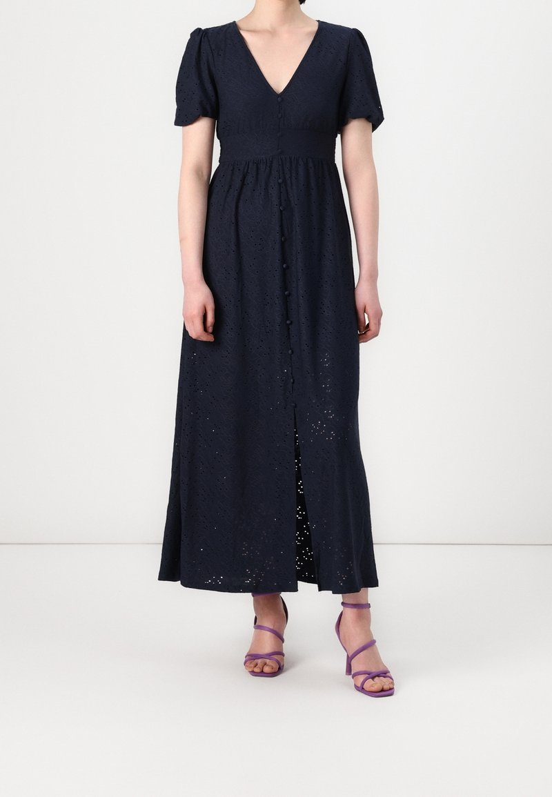 Femme portant une robe maxi bleu marine œillet avec manches courtes et boutons à l'avant, associée à des sandales à talons pourpres à lanières.