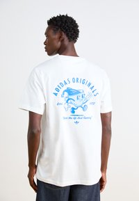 Λευκό βαμβακερό μπλουζάκι με μπλε γραφικό σχέδιο στην πλάτη, που περιλαμβάνει το κείμενο "ADIDAS ORIGINALS" και έναν χαρακτήρα κινούμενο σε πόζα τρεξίματος.