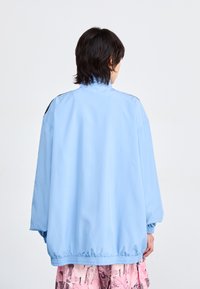 Chaqueta cortaviento de gran tamaño en azul claro, con un alto cuello, puños y dobladillo elásticos. Textura suave, hardware mínimo y diseño simple.
