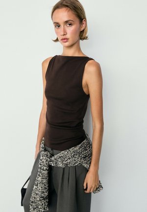 Jeune femme portant un haut sans manches sombre et un pantalon gris avec un pull tricoté noir et blanc noué autour de la taille, devant un mur uni.