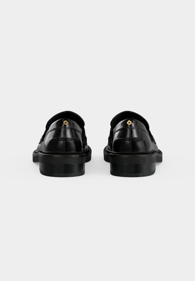 Paire de chaussures de ville en cuir noir poli vues de dos, chacune avec un petit emblème de fleur dorée près du talon.