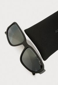 Occhiali da sole neri con una forma rettangolare elegante, lenti grigie e opache, poggiati accanto a una custodia morbida nera.