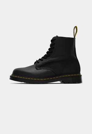 Zwarte leren veterschoen tot de enkel met gele stiksels op de zool en een merklabel aan de hak, zijaanzicht op een effen achtergrond.