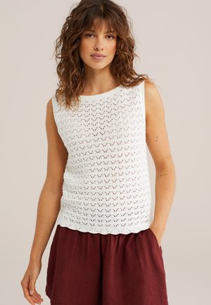 Frau mit lockigem Haar, die ein ärmelloses, weißes, gestricktes Oberteil und weite, kastanienrote Shorts trägt, stehend mit einer Hand in der Tasche.