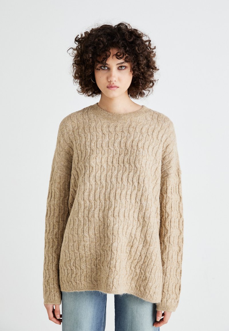 Beige kabelstrikket genser med lange ermer, vid passform og ribbestrikket rund hals. Myk tekstur, kombinert med lyseblå jeans.