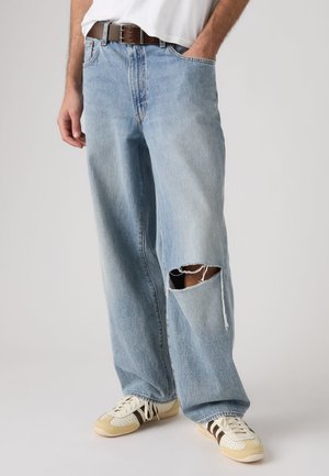 Man wearing light blue ripped jeans with a tear on the right knee, white sneakers with beige and black stripes, and a brown belt.