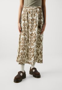 Falda larga en crema con patrones florales marrones, con una cintura elástica y lazos. Combinada con zapatos marrones y calcetines claros.