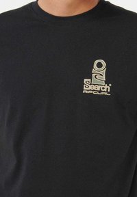 Sort langærmet T-shirt lavet af bomuld, med et lille beige grafisk logo på venstre brystområde. Enkel rund halsudskæring.