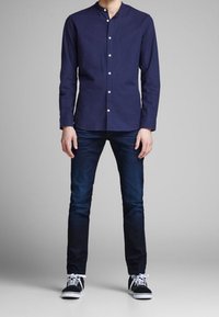 Camicia a maniche lunghe blu navy con piccoli bottoni, abbinata a jeans slim fit blu scuro e sneakers nere con suole bianche. Design semplice, senza motivi.