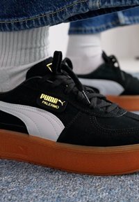 Adidași din suede negru cu accente albe și logo auriu. Prezintă o talpă din cauciuc texturată și un design cu șireturi. Sosete albe cu nervuri vizibile.