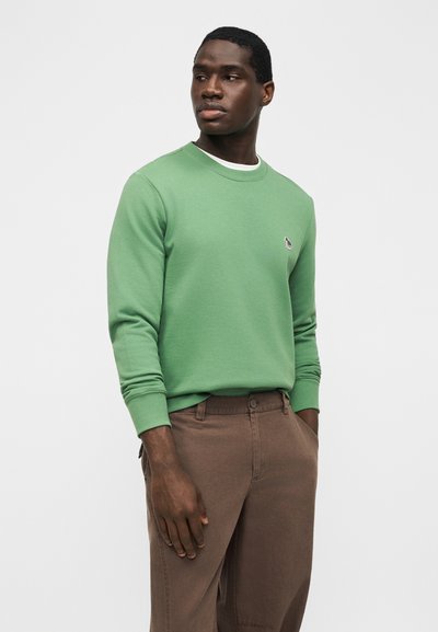 Άνδρας με ανοιχτό πράσινο φούτερ και καφέ παντελόνι, στέκεται με τα χέρια στις τσέπες, κοιτάζοντας προς το πλάι μπροστά σε απλό γκρι φόντο.