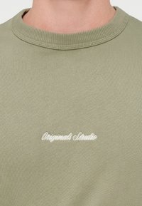 Λαδί πράσινο μπλουζάκι με ριμπ λαιμόκοψη και απαλή υφή; έχει λευκό κεντημένο κείμενο «Originals Studio» στο στήθος.