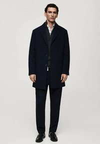 Cappotto di lana navy con colletto a rever, chiusura con un bottone, sovrapposto a un maglione con zip sul collo e pantaloni coordinati, abbinato a scarpe nere.