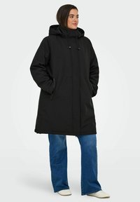 Svart huvad parka med avslappnad passform, dragkedjeförsedda fickor och justerbara dragsnören. Bärs över utsvängda jeans och vita skor.