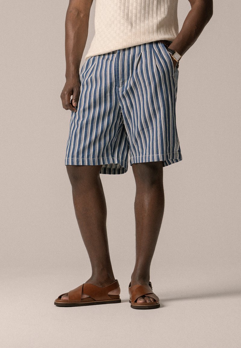 Blaue und weiße gestreifte Shorts aus leichtem Stoff, mit einem elastischen Bund und einer lässigen Passform. Kombiniert mit braunen Ledersandalen.