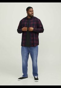 Ruitjes flanellen shirt in donkerrood en marineblauw, met knoopjes op de kraag, gedragen over een zwart T-shirt. Gecombineerd met lichtblauwe jeans en zwarte sneakers.