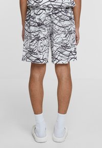 White shorts with a black abstract line pattern, featuring an elastic waistband and pockets. Textured fabric, knee-length design.