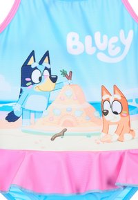 Maillot de bain une pièce avec un fond bleu clair et un ourlet froncé rose, représentant des chiens animés construisant un château de sable.