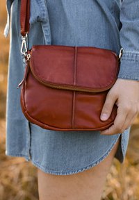 Ruda odinė crossbody rankinė su užtrauktuku, apvalios formos ir priekinio dangtelio. Ji turi diržą su metalinėmis detalių akcentais.