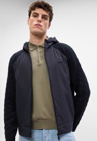 Casaco preto com fecho e capuz, apresentando mangas em malha texturizada. Usado sobre uma camisa polo verde-claro, com jeans azul na cintura.