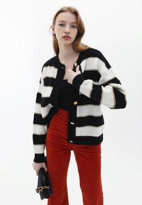 Svart och vit randig cardigan med ribbad struktur, prydd med knappar i guldton, kombinerad med oranga manchesterbyxor och en svart clutch-väska.