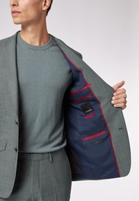 Grijze colbert met een gladde textuur, voorzien van een marineblauwe voering met rode accenten en meerdere zakken. Draagt men over een grijze trui.