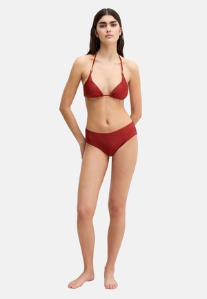 Kobieta stojąca boso, ubrana w czerwony bikini związywany na szyi z drewnianymi koralikami oraz pasujące wysokie majtki bikini, na jednolitym tle.