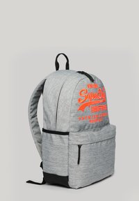 Mochila de tela gris con correas de hombro acolchadas, presenta un logo naranja brillante, bolsillo frontal y un acento negro en la parte inferior. Cierre de cremallera.