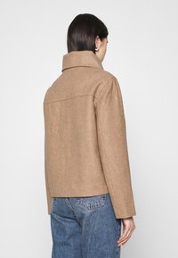 Brun uld-blandingsjakke med høj krave, et struktureret fit og lange ærmer. Bagfra ses sømdetaljer og tekstur. Båret sammen med blå jeans.