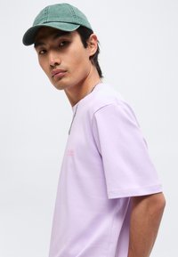 Νέος άνδρας με πράσινο καπέλο και ανοιχτό μοβ T-shirt, σε προφίλ με ουδέτερη έκφραση σε απλό λευκό φόντο.