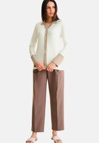 Geribbeld wit blouse met beige kraag en manchetten, gecombineerd met losse bruine broeken. Het model draagt witte schuifslippers.
