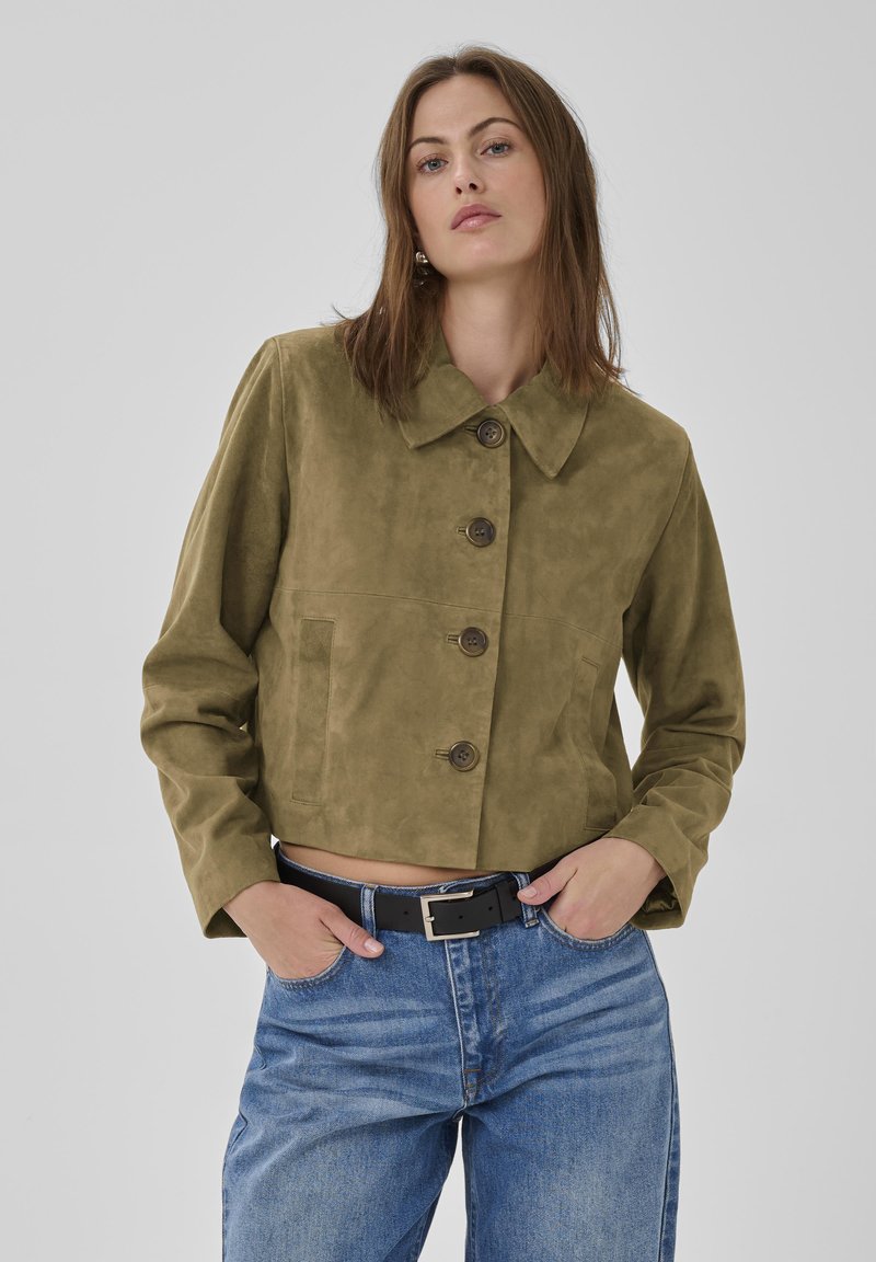 Vrouw die een cropped olijfgroene suède jas met grote knopen draagt, blauwe high-waist jeans en een zwarte riem, staand met handen in de zakken.