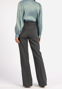 Personne portant un chemisier en satin bleu sarcelle à manches longues et un pantalon large taille haute gris foncé, debout de dos face au spectateur.