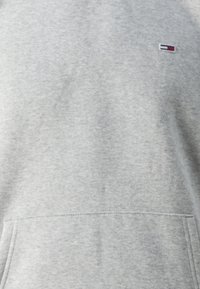 Sudadera gris con textura suave, que cuenta con un bolsillo tipo canguro en la parte delantera y un pequeño logo en azul, rojo y blanco en la parte superior izquierda.
