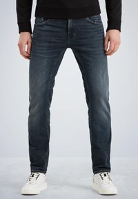 Jeans en denim bleu avec une coupe slim, présentant un design classique à cinq poches. Le tissu a un aspect usé et est associé à des baskets blanches.