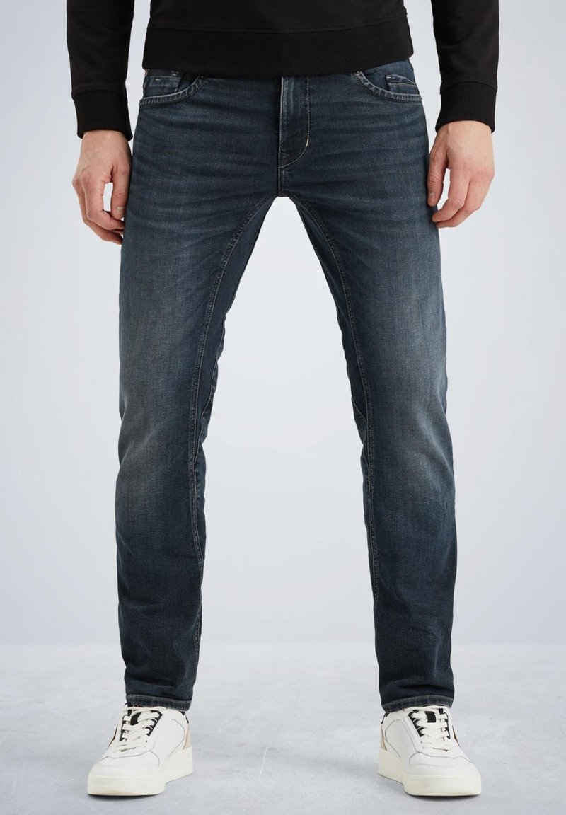 Jeans en denim bleu avec une coupe slim, présentant un design classique à cinq poches. Le tissu a un aspect usé et est associé à des baskets blanches.