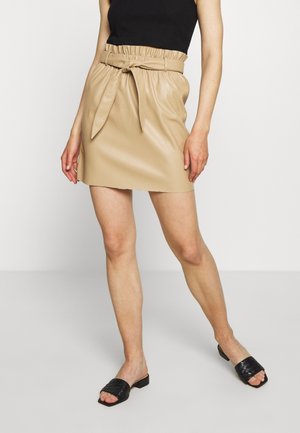 Beige Kunstleder-Minikleid mit gerafftem Bund und Bindegürtel, kombiniert mit schwarzen High-Heel-Pantoletten.