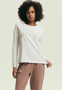 Biała koszulka z długim rękawem o luźnym kroju zdobiona jasnobrązowym nadrukiem. Połączona z brązowymi, luźnymi spodniami.