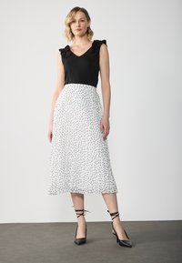 Schwarzes Oberteil mit Rüschensleeves, kombiniert mit einem weißen Midirock, der schwarze Punkte hat. Schwarze, spitze Absatzschuhe mit Knöchelriemen.
