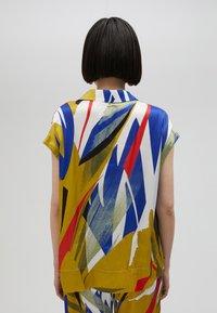 Blúzka s krátkymi rukávmi s farebným geometrickým vzorom v žltých, modrých, červených a zelených farbách. Hladká textúra, golier s gombíkmi a voľný strih.