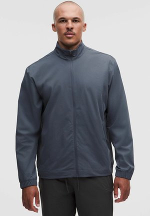 Uomo con i capelli corti che indossa una giacca grigio scuro con zip e pantaloni neri, in piedi davanti a uno sfondo chiaro e semplice.