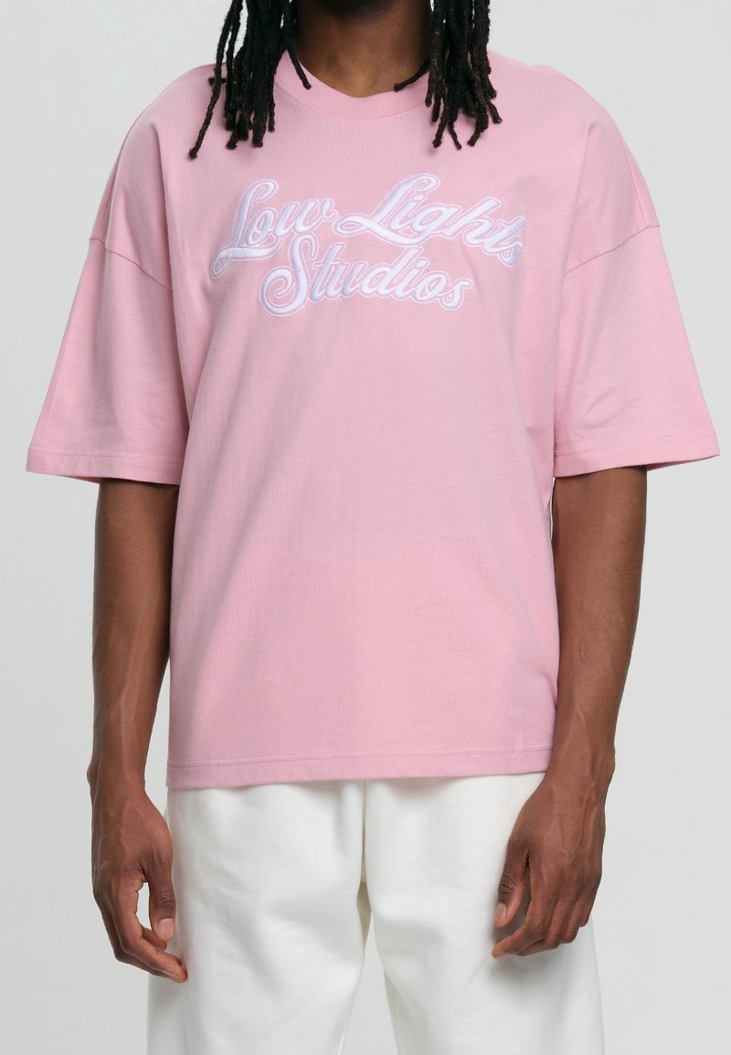 Homme portant un t-shirt rose ample avec le texte "Low Lights Studios" en cursive blanche, accompagné d'un pantalon blanc, se tenant devant un fond uni.